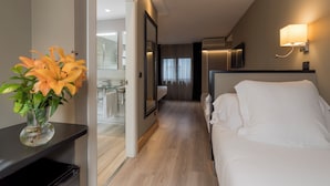 Una camera d'albergo con un letto, un comodino, un vaso con fiori, uno specchio e un bagno visibile attraverso una porta aperta.
