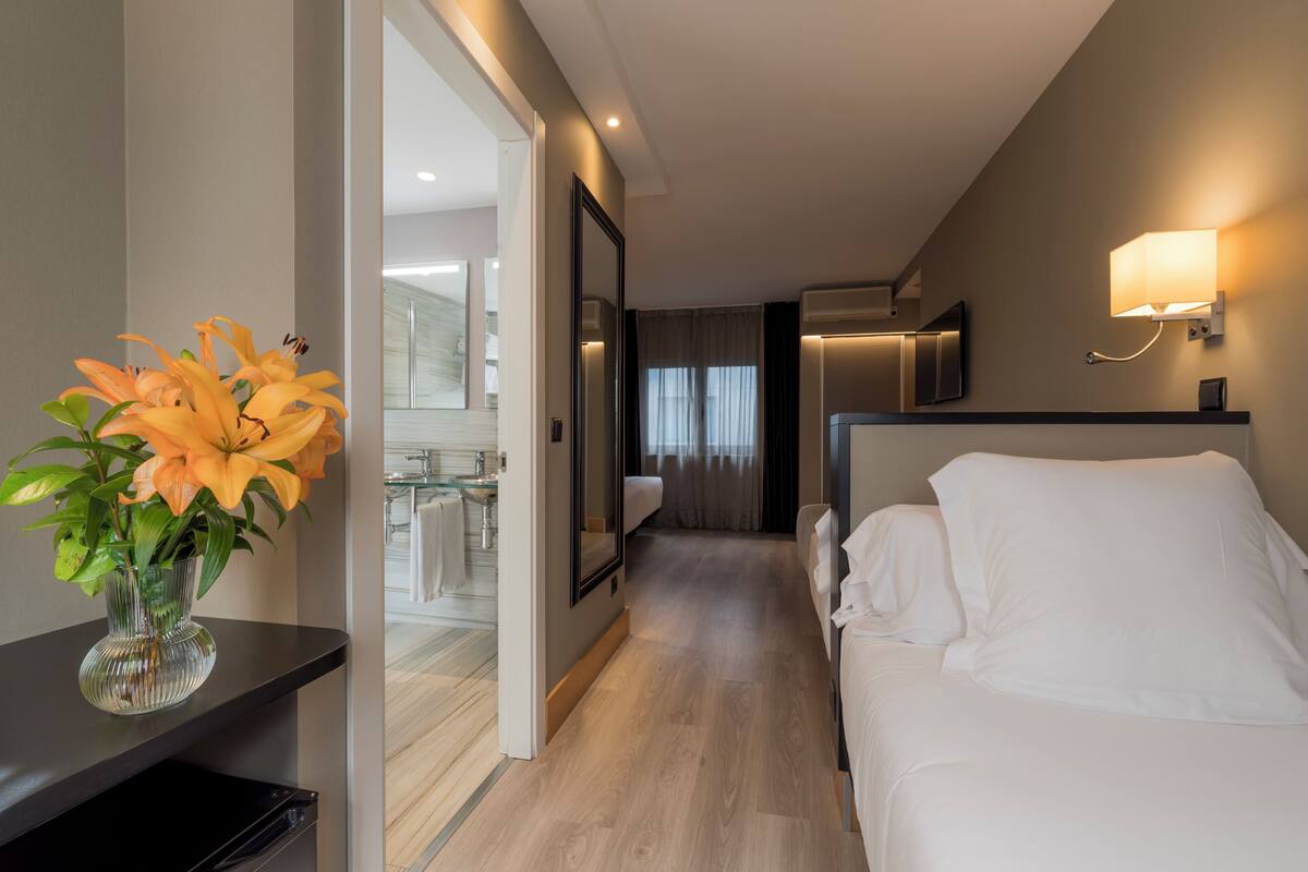 A hotel room with a bed, a nightstand, a vase with flowers, a mirror, and a bathroom visible through an open door.