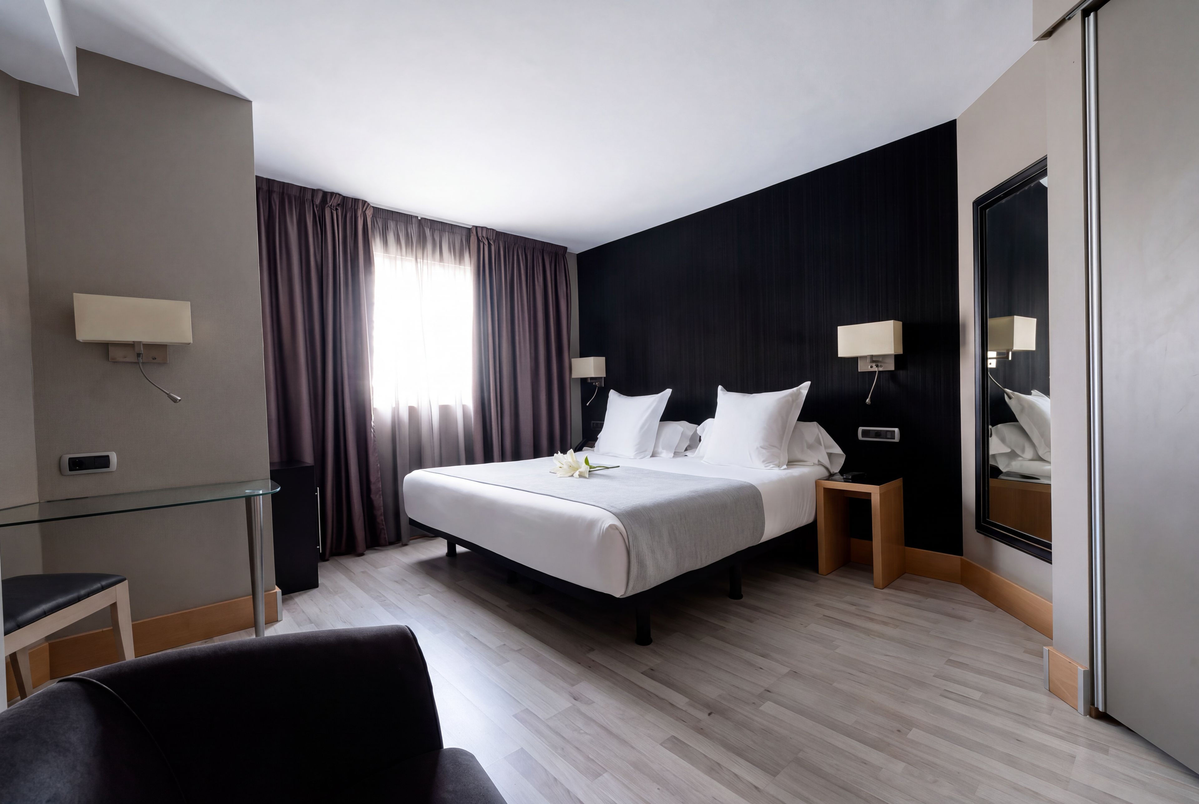 A hotel room with a bed, bedside tables, a desk, a chair, a mirror, and a window with curtains.