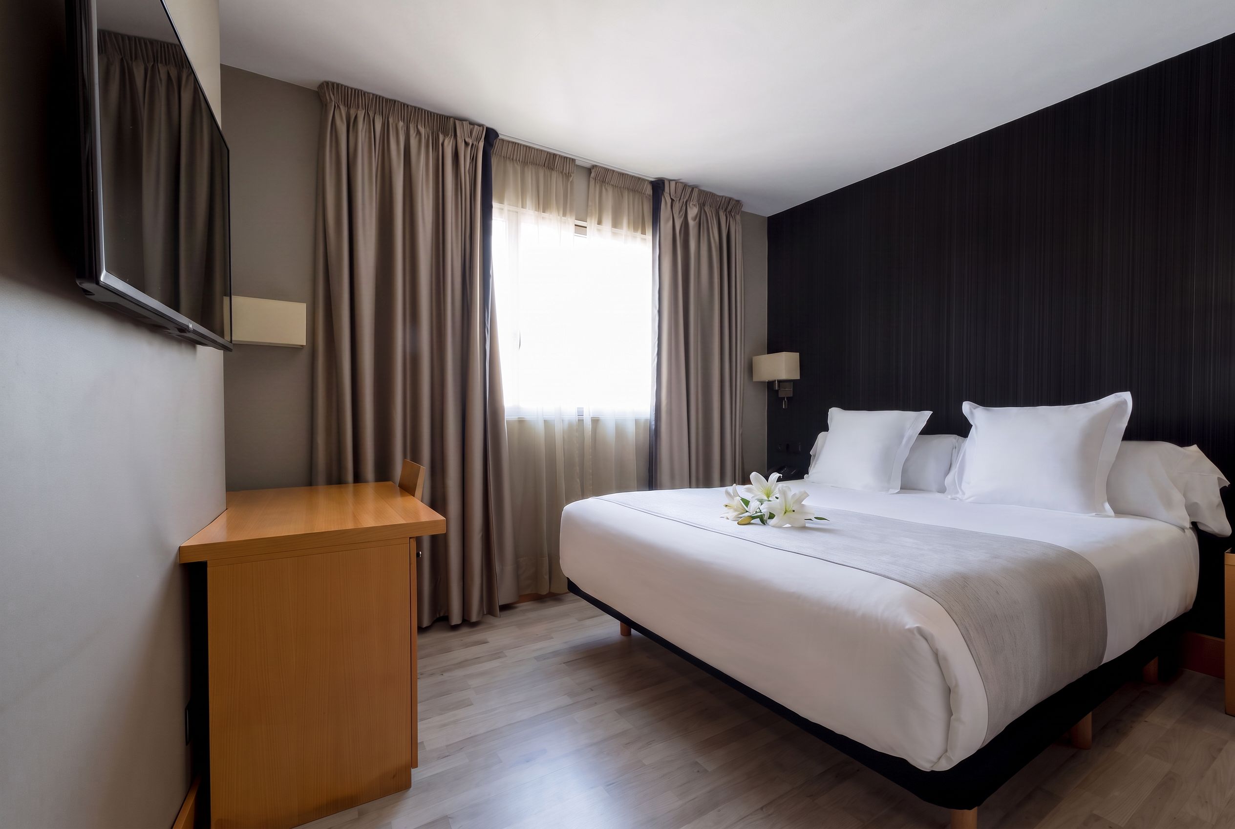 A hotel room with a bed, a desk, a mirror, and a window with curtains.