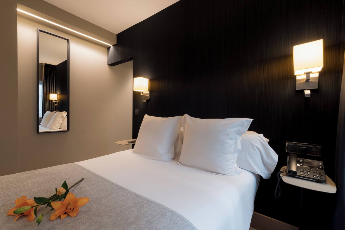 A hotel room with a large bed, a bedside table with a phone, a mirror, and a wall-mounted light fixture.
