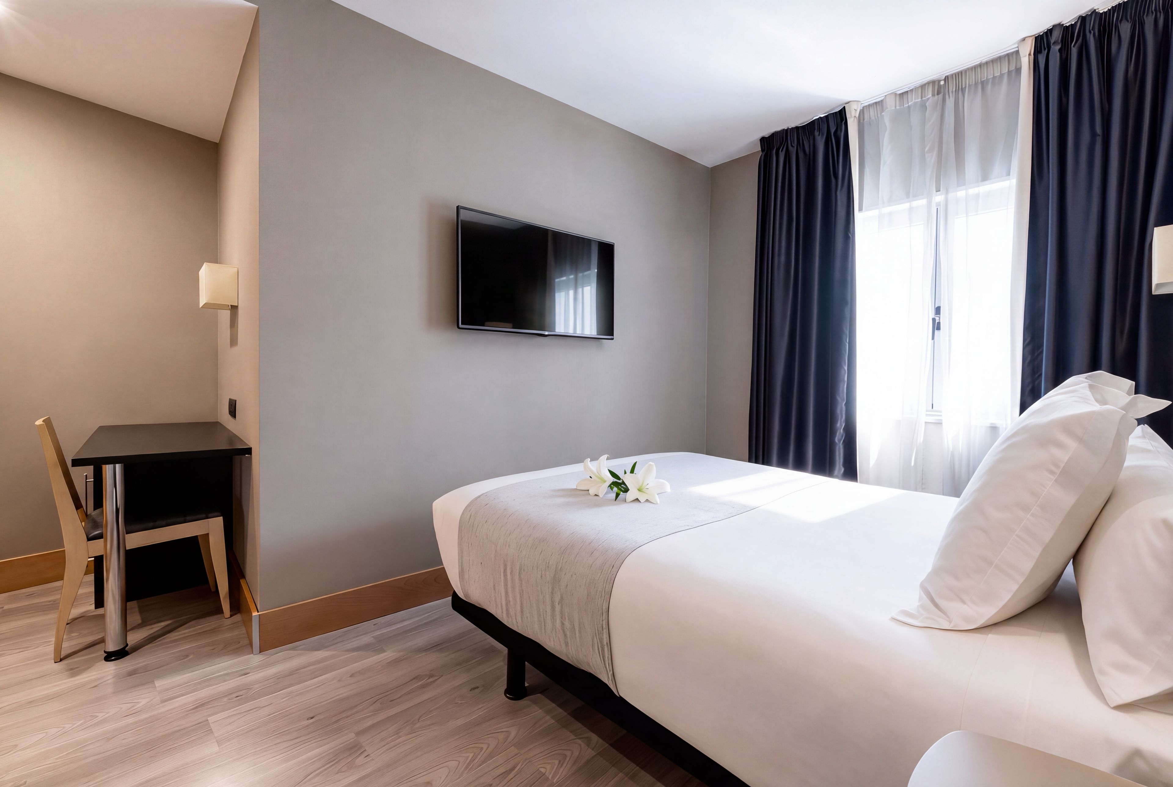Habitación de hotel con cama, escritorio, silla, televisor y ventana con cortinas.