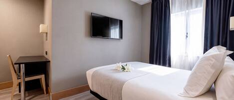 Habitación de hotel con cama, escritorio, silla, televisor y ventana con cortinas.