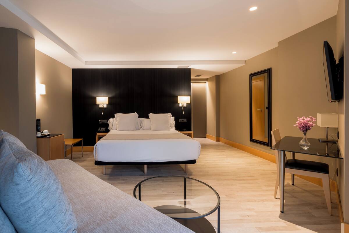 A modern hotel room with a large bed, bedside tables, a TV, and a small table with a vase.