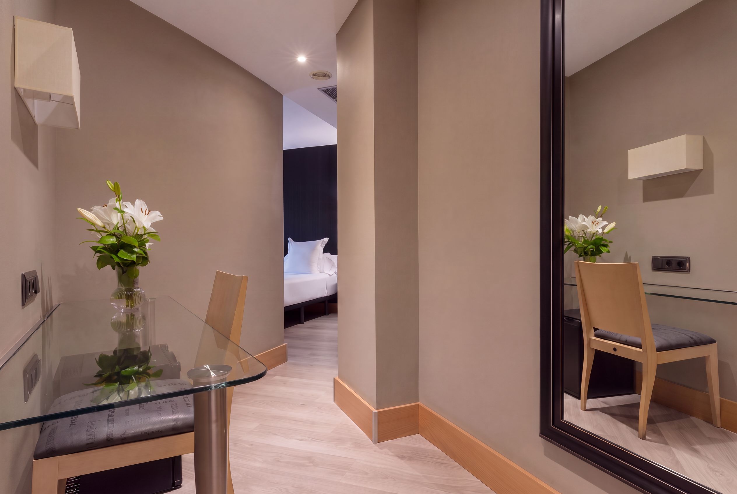 A hotel room with a glass table, two chairs, a large mirror, and a bed with white bedding.