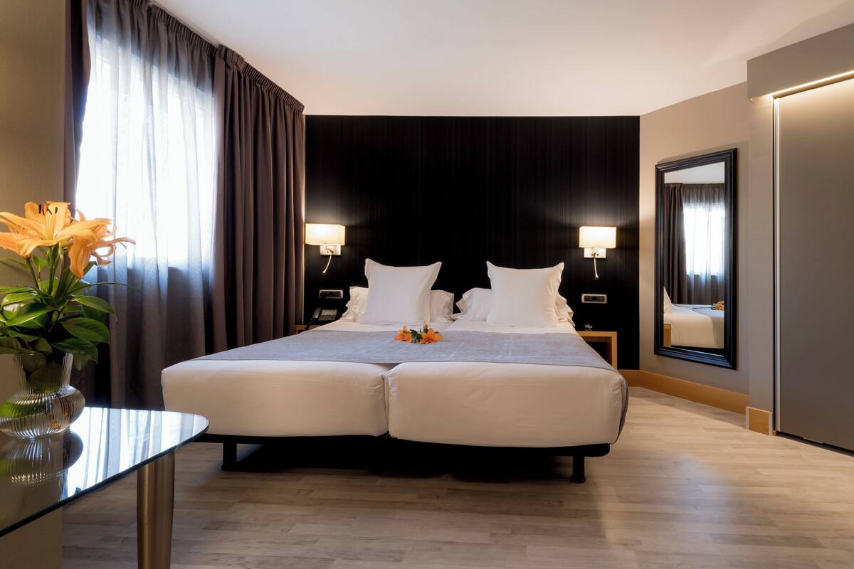 A double bed with white linens, two bedside tables with lamps, a large mirror, and a vase with flowers on a glass table.