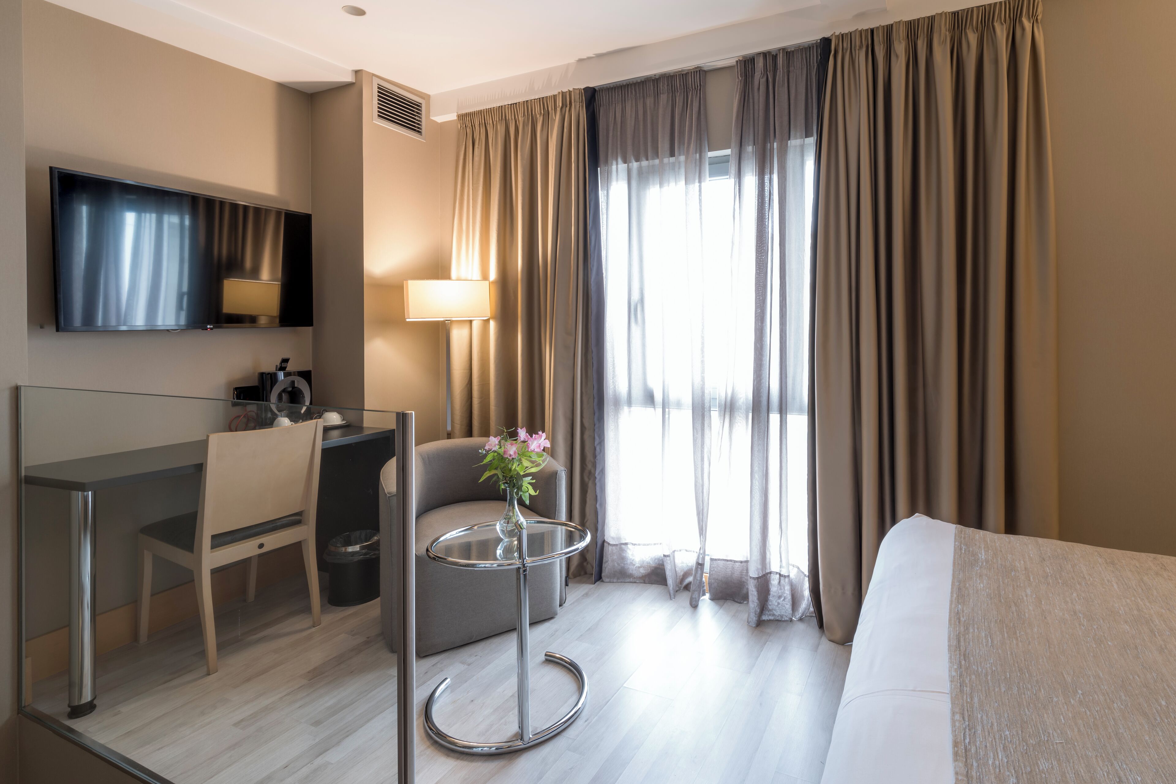 A modern hotel room with a flat-screen TV, a desk with a chair, a small table with a vase, and a bed.