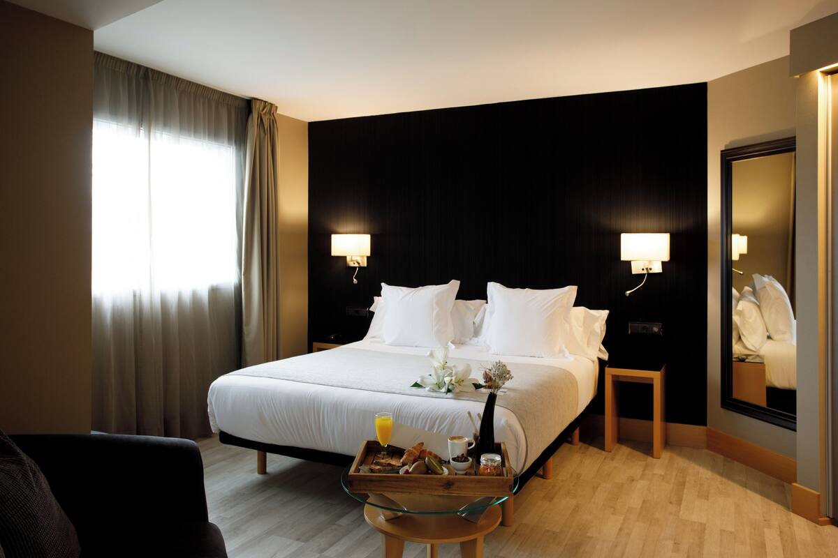 A hotel room with a large bed, two bedside tables with lamps, a small wooden stool, and a tray with breakfast items.