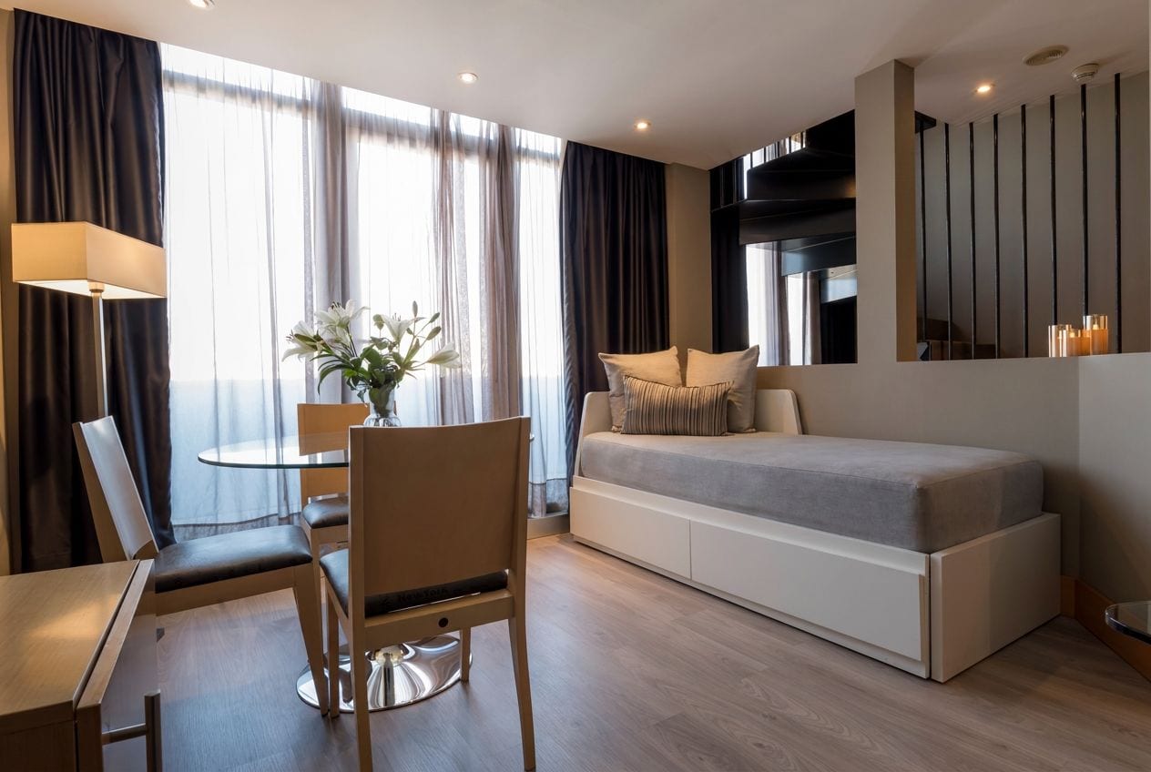A modern hotel room with a sofa bed, a dining table with chairs, a vase with flowers, and a large window with curtains.