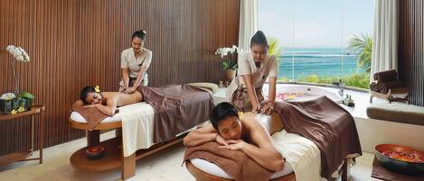 A spa room with two massage tables, a large window with a view of the ocean, and a wooden wall.