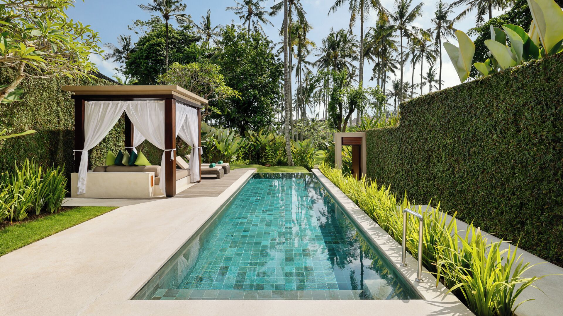 A rectangular swimming pool with clear blue water, surrounded by a hedge, a wooden deck, and a covered seating area with cushions.