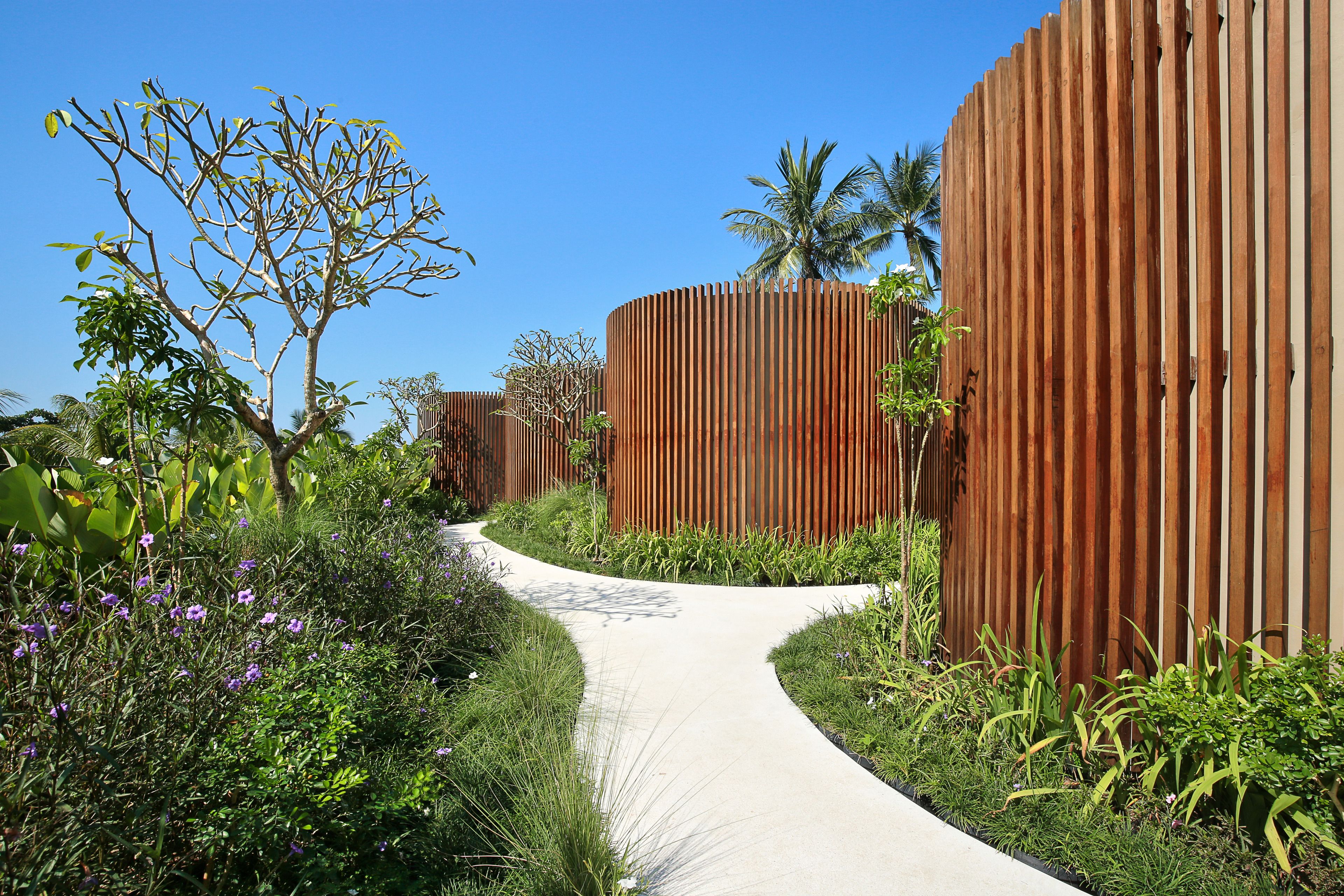 A pathway flanked by tall wooden fences, surrounded by lush greenery and flowering plants.