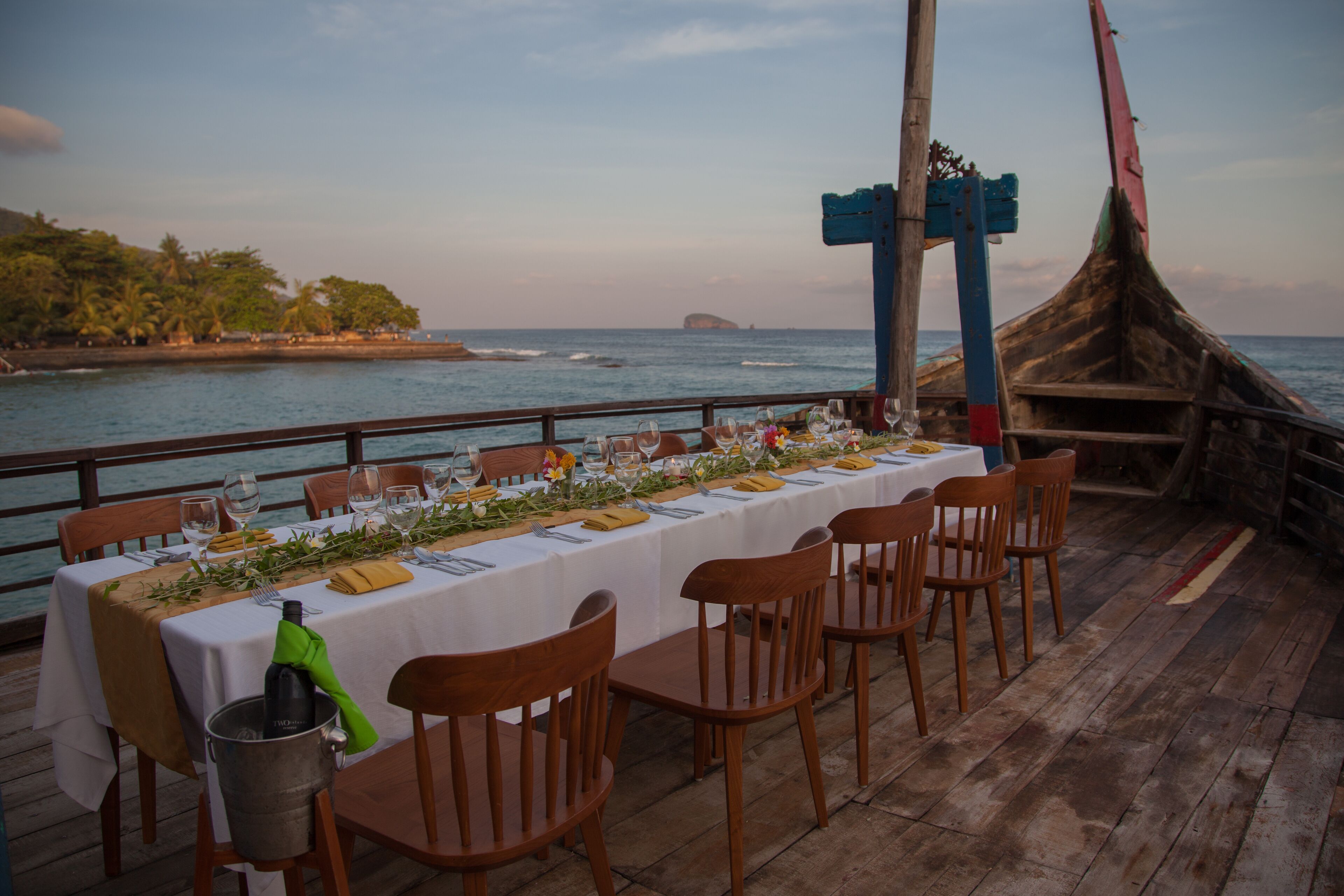 A long dining table set for a meal with chairs, a view of the ocean, and a traditional boat structure in the background.