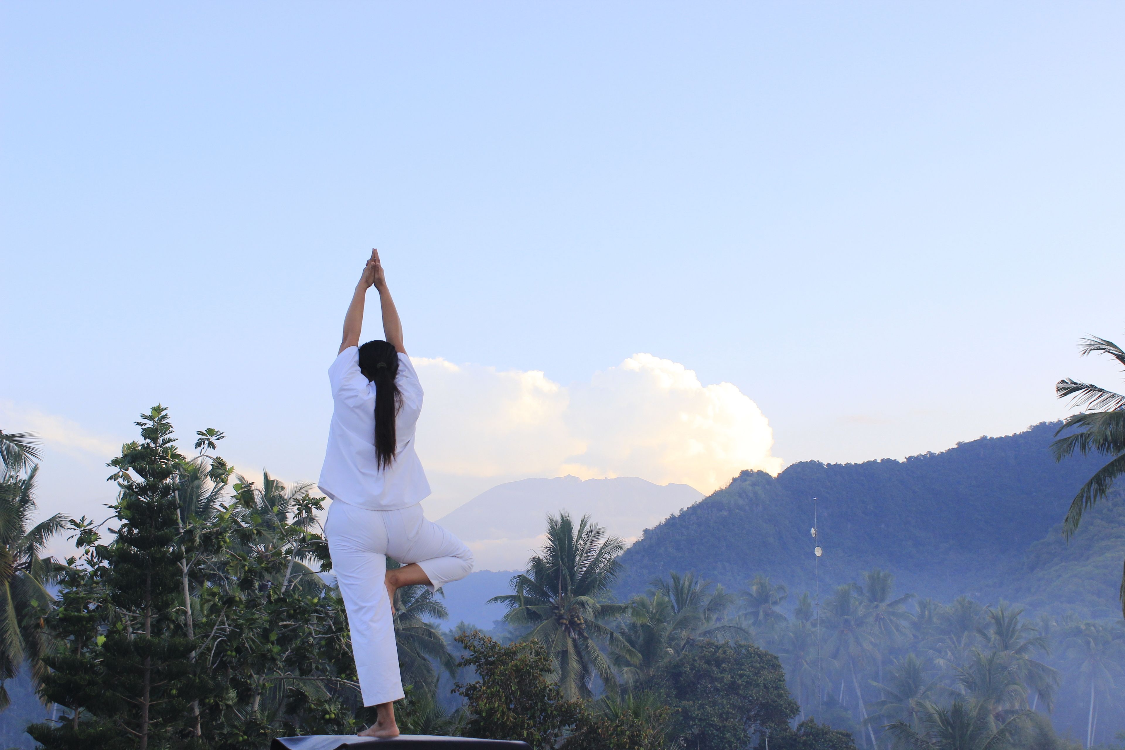 Una persona realizando una postura de yoga sobre una plataforma, con un paisaje montañoso y palmeras al fondo.