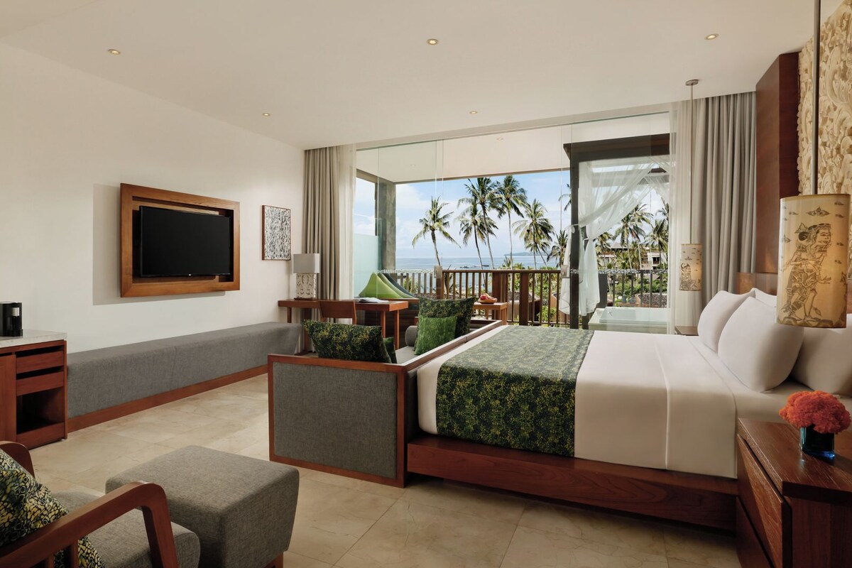 Habitación de hotel con cama, televisión, escritorio, silla y balcón con vistas a palmeras y al océano.