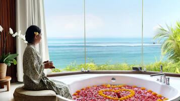 A person sitting by a large bathtub filled with flower petals, overlooking a scenic ocean view.