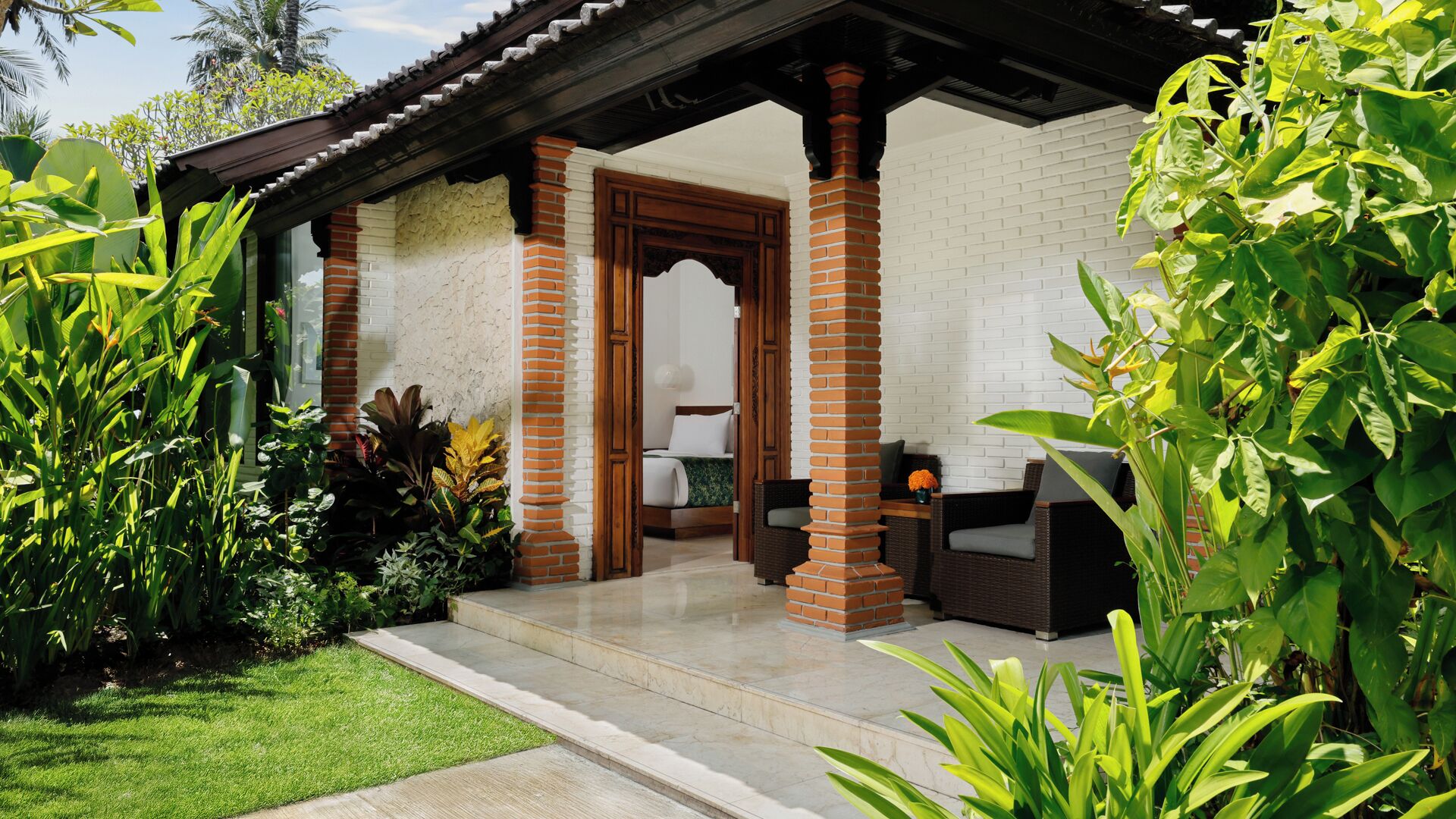 A covered outdoor seating area with a brick wall, a wooden door, and a bed visible inside.