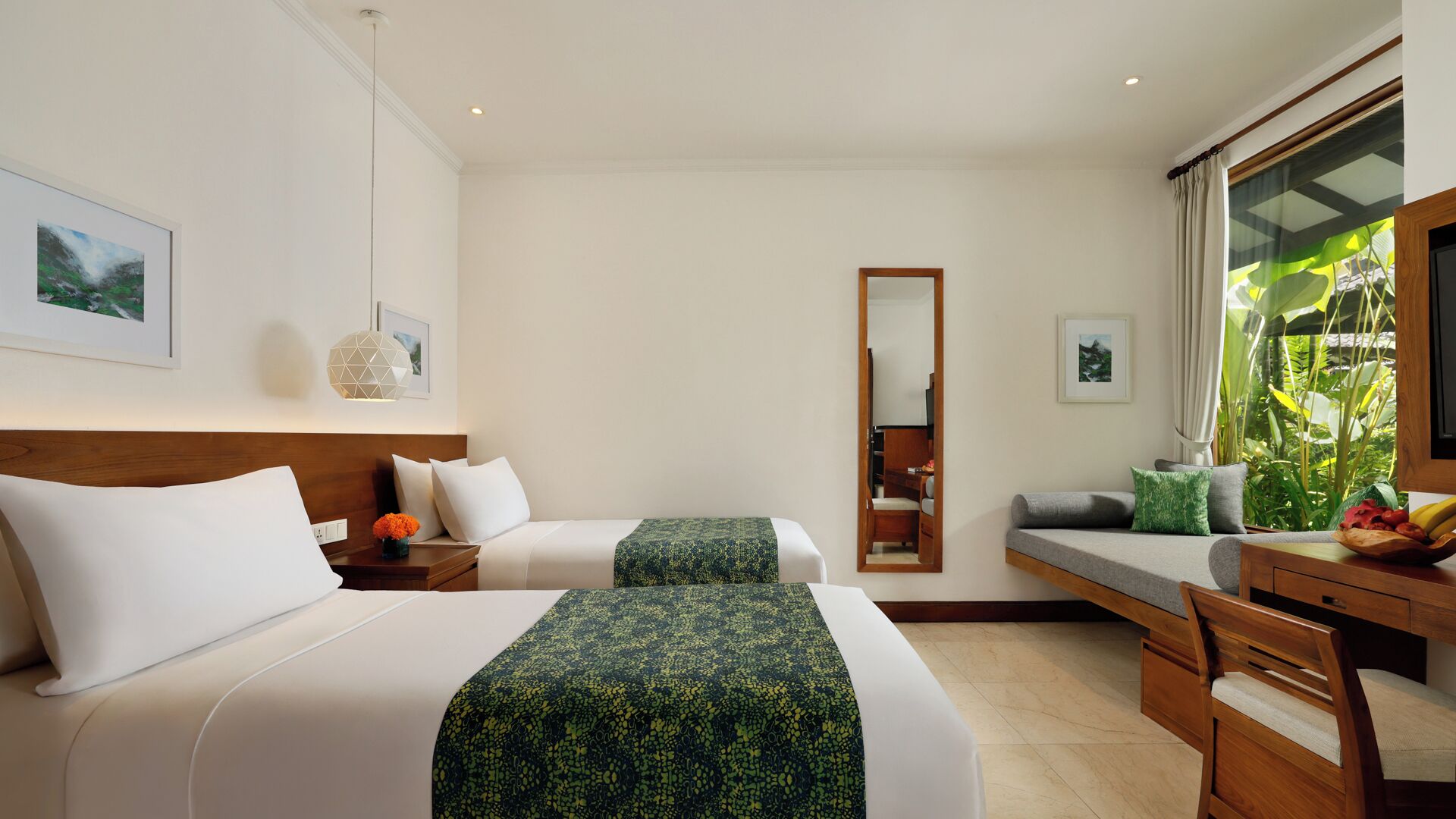 A hotel room with two beds, a desk, a mirror, and a window with a view of greenery.