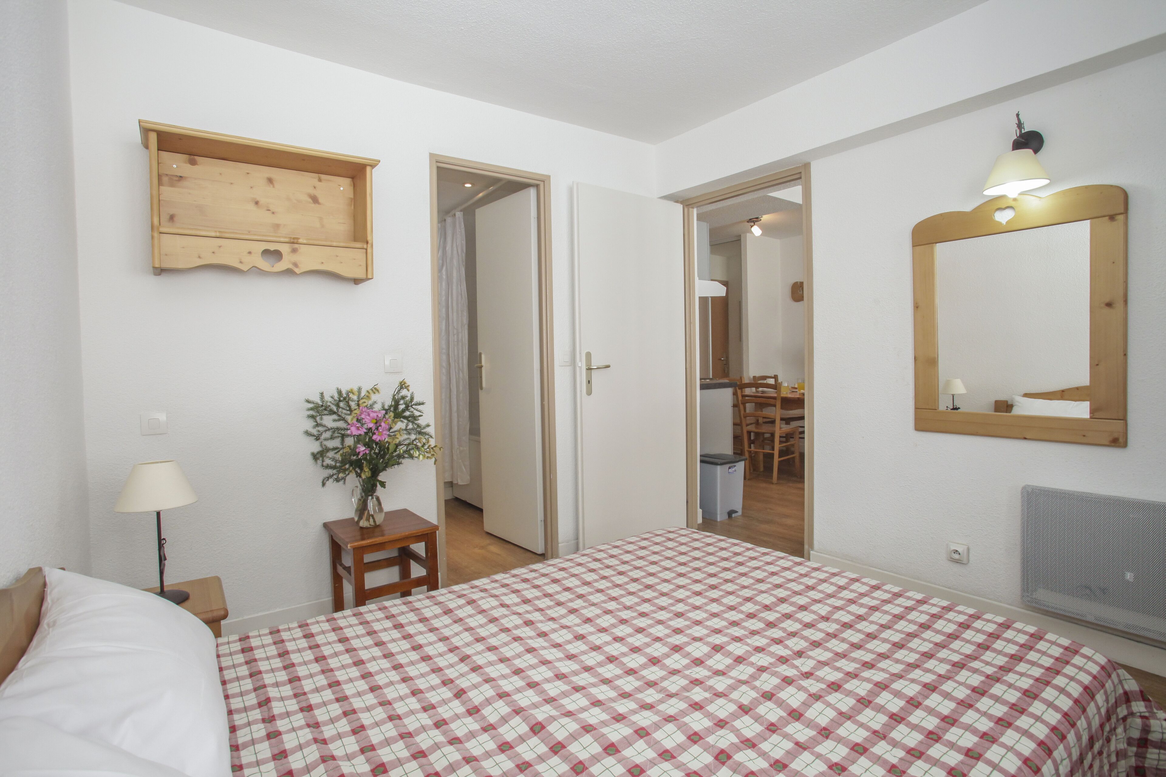 A bedroom with a checkered bedspread, a wooden shelf, a mirror, and a small table with a lamp and flowers.