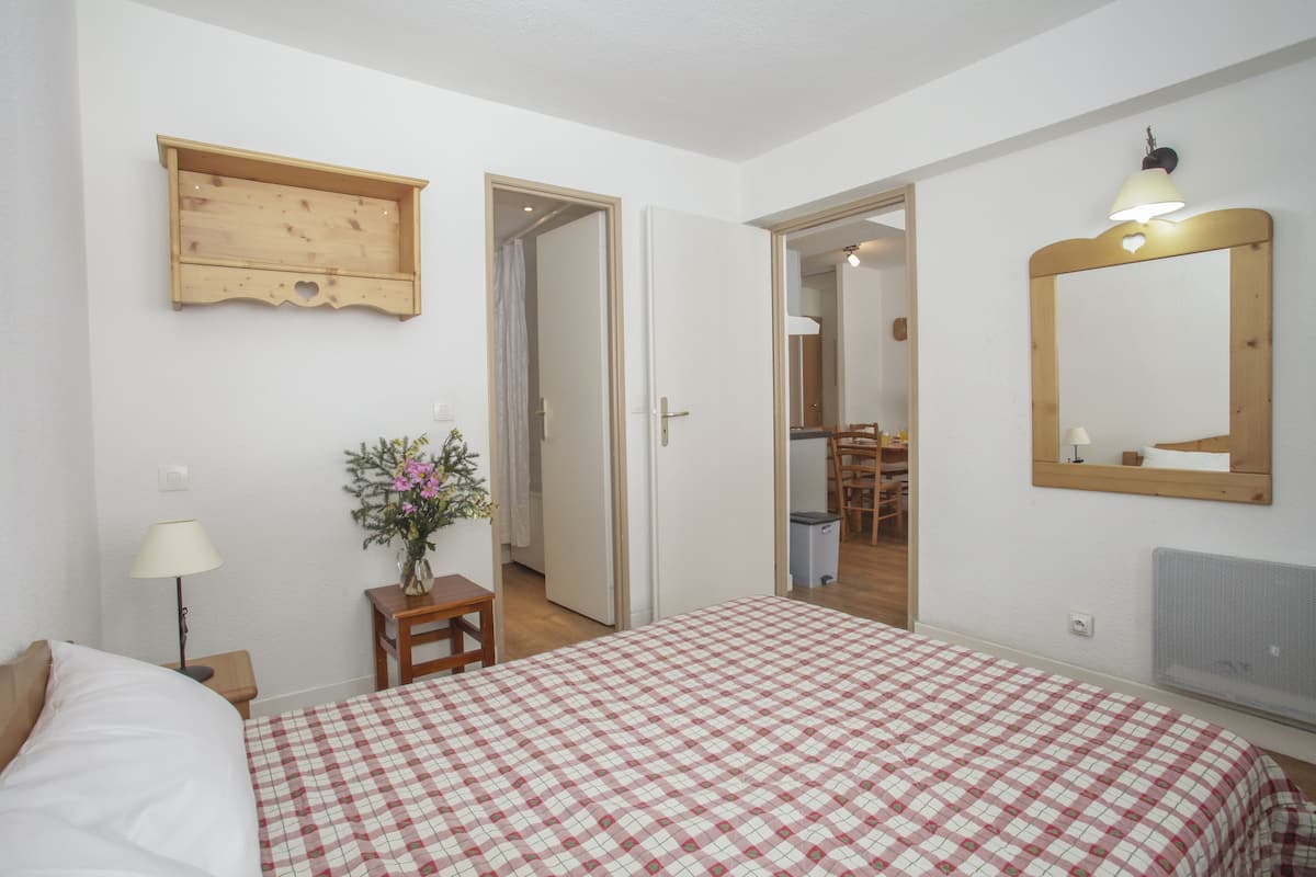 A bedroom with a checkered bedspread, a wooden shelf, a mirror, and a small table with a lamp and flowers.