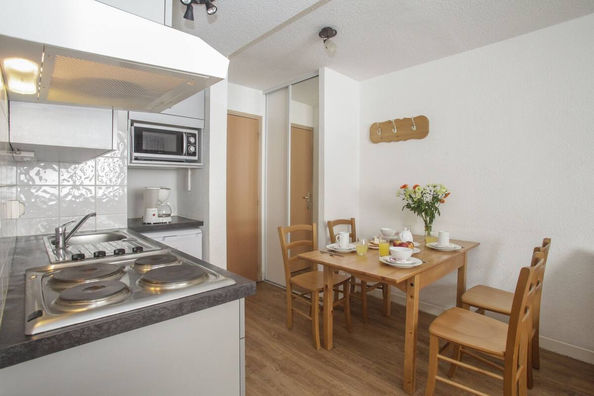 A compact kitchen with a stove, microwave, and sink. There is a dining table with chairs and a vase of flowers.