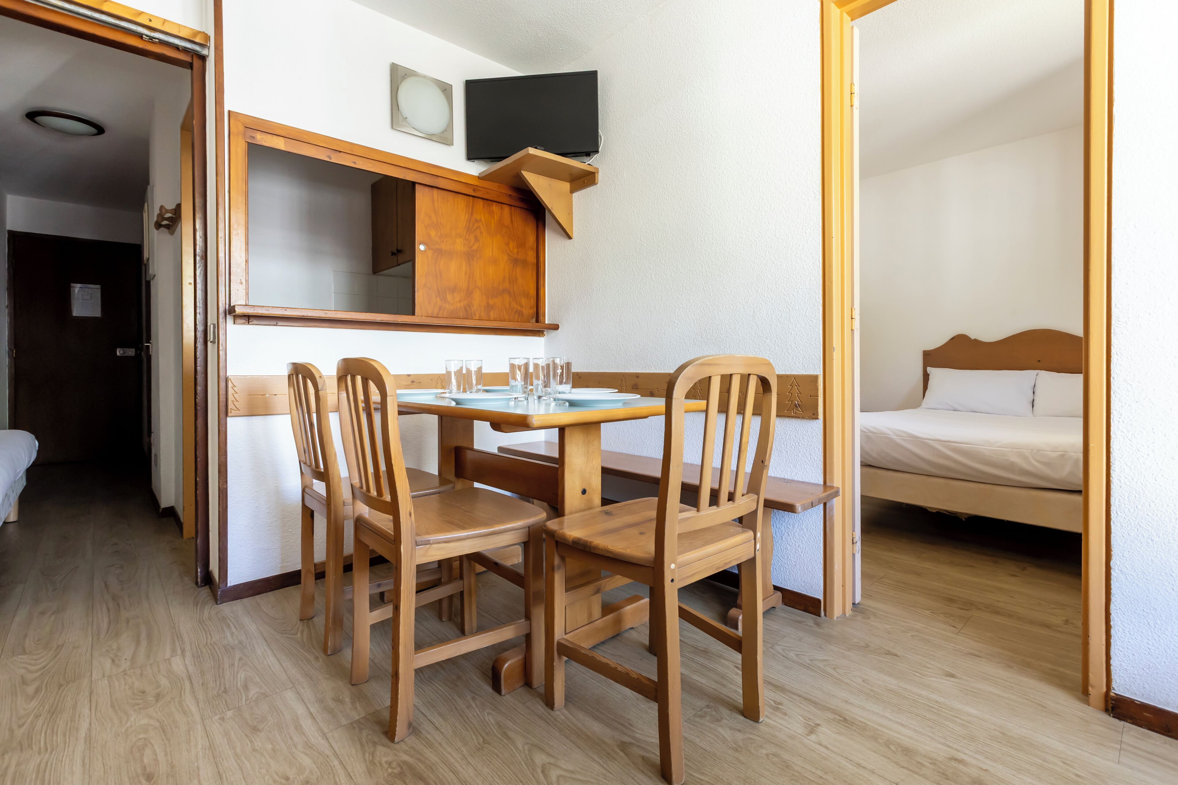 A room with a wooden table, four chairs, a bed, a television, and a mirror.