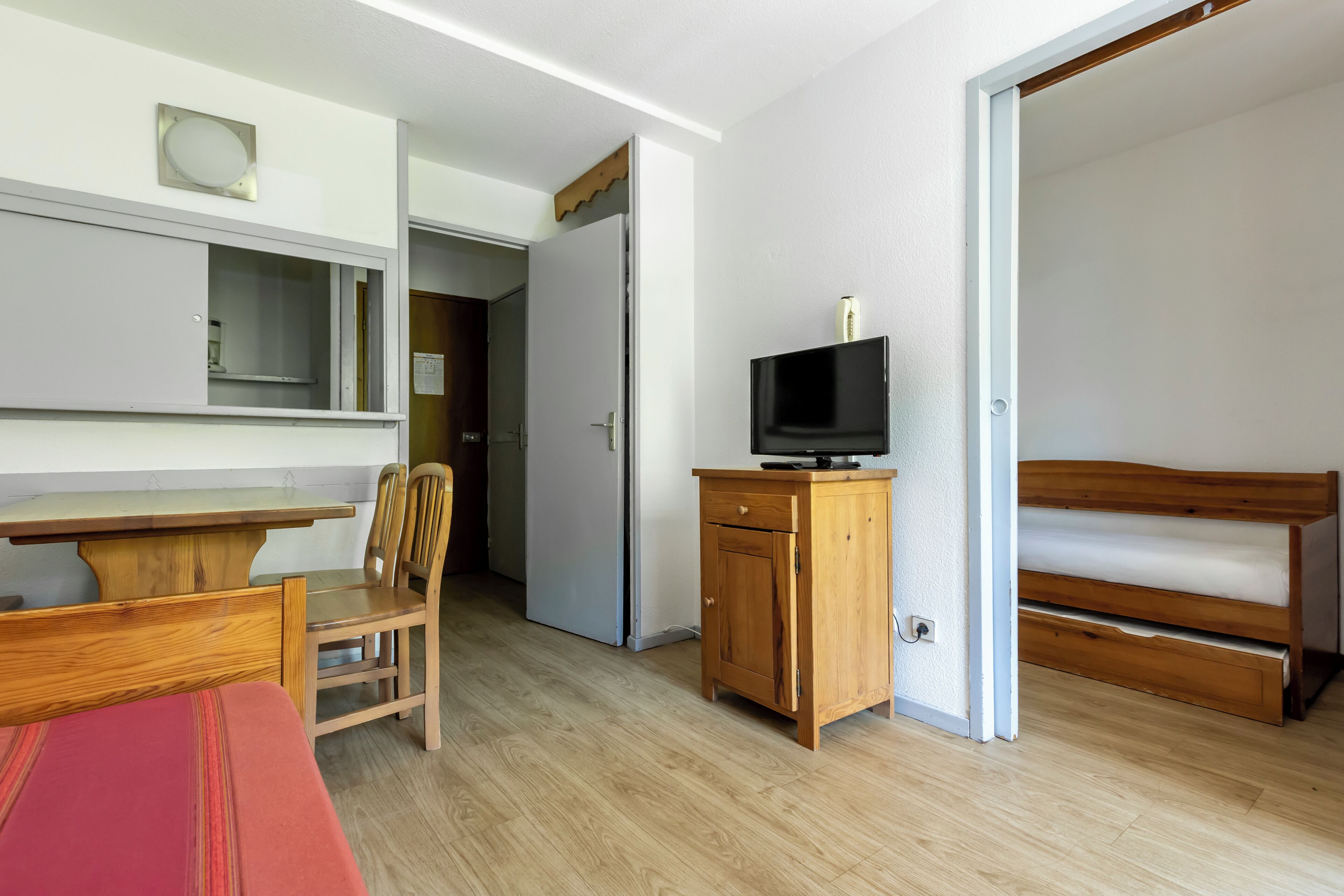Una habitación compacta con una cama de madera, un televisor sobre un mueble de madera, una mesita con una silla y una puerta que da a otra habitación.