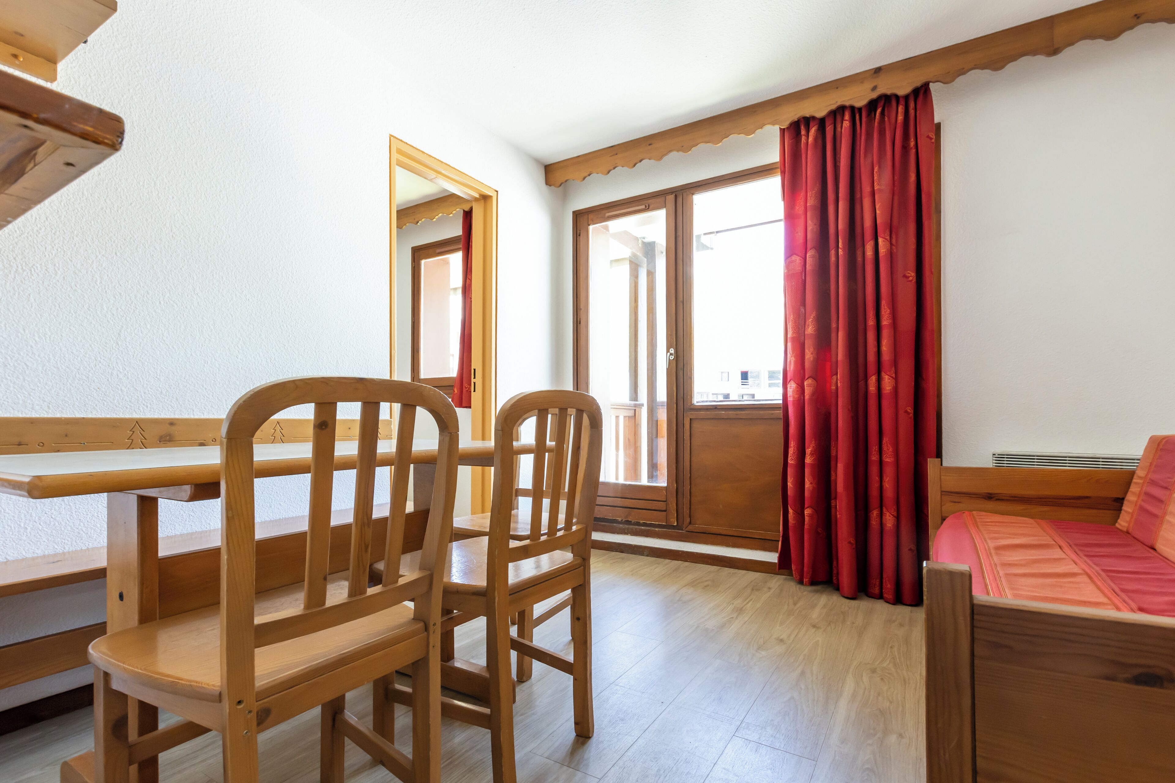 A room with a wooden table, two chairs, a bed, and a window with red curtains.