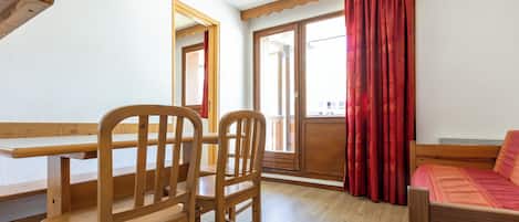 A room with a wooden table, two chairs, a bed, and a window with red curtains.