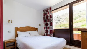 A bedroom with a wooden headboard, a single bed, a nightstand, a window with curtains, and a view of a hillside. - Odalys Residence le hameau du Borsat (Tignes)