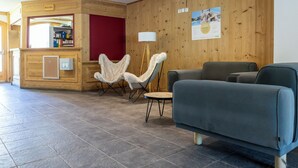 A reception area with a wooden wall, a reception desk, a blue sofa, and a white furry chair. - Odalys Residence le hameau du Borsat (Tignes)