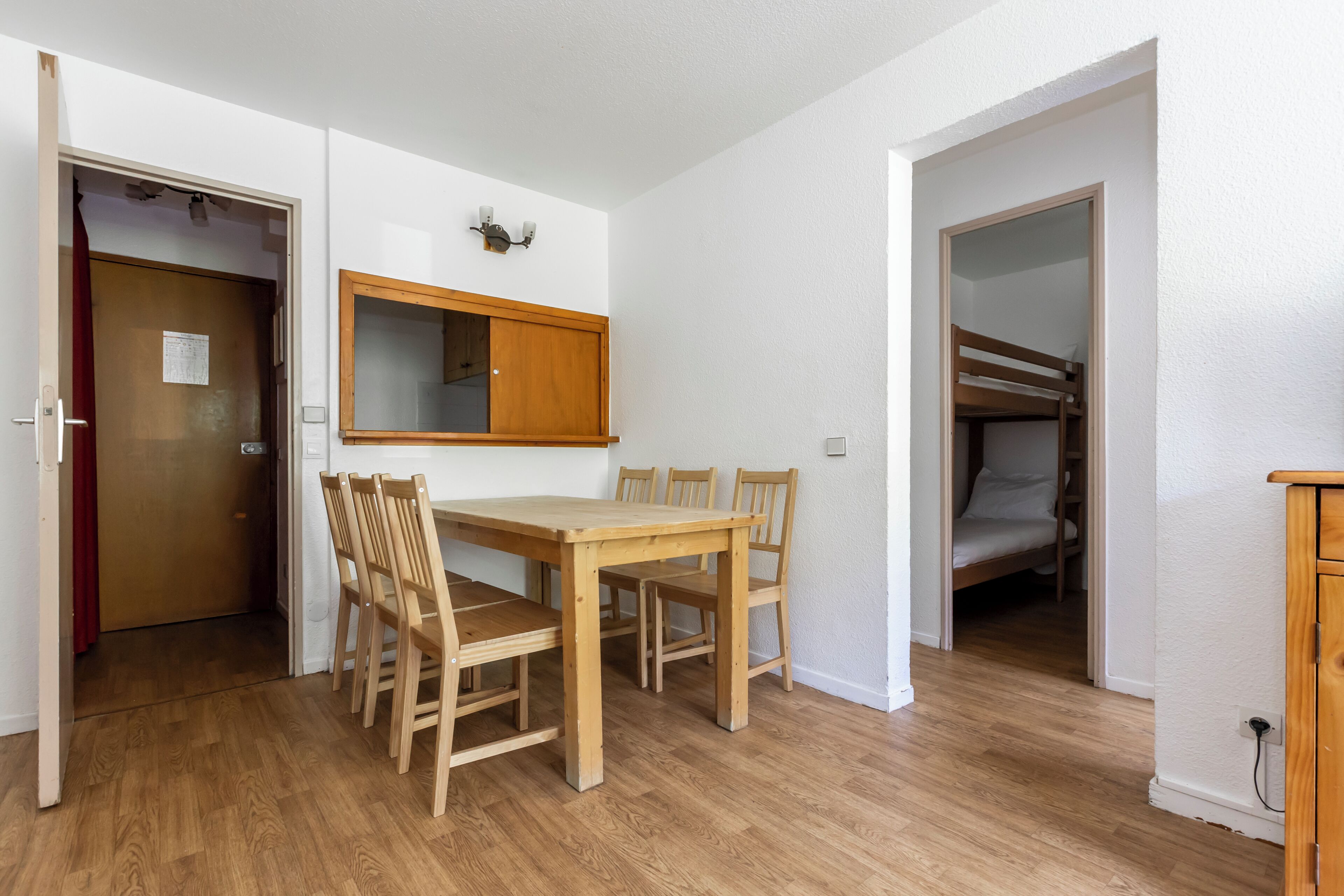 A room with a wooden dining table and chairs, a wooden cabinet, and a bunk bed in the adjacent room.