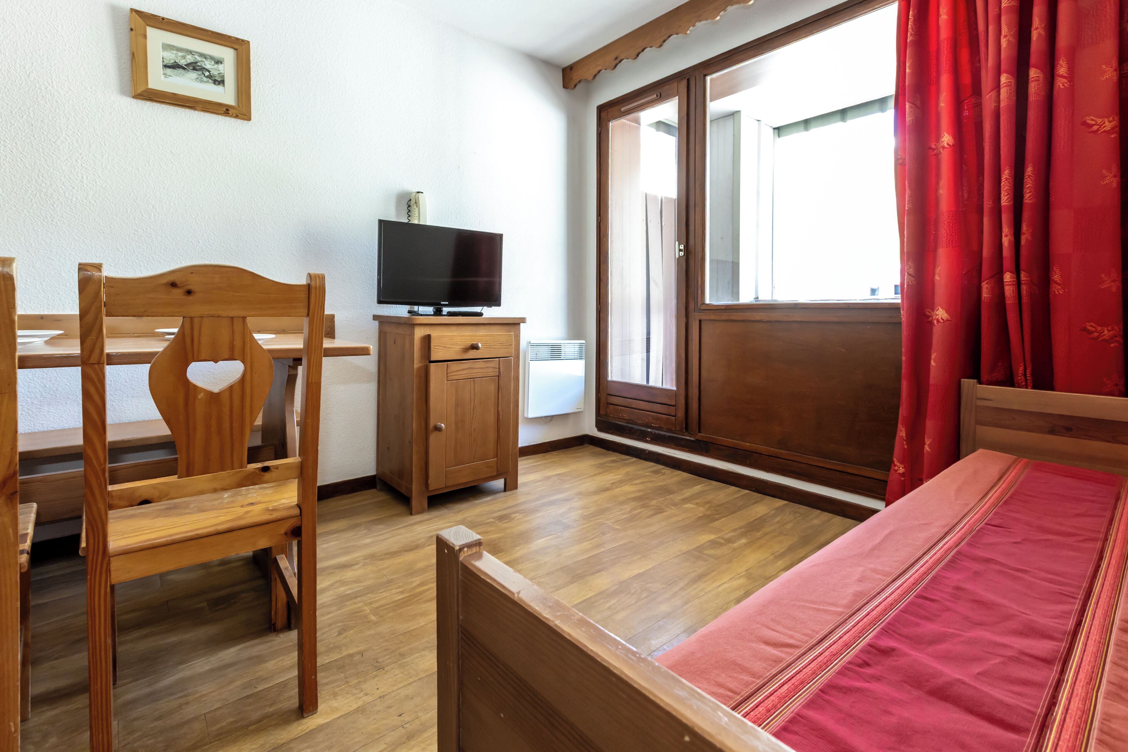 Una habitación con cama, una silla de madera, un televisor sobre un mueble y una ventana con cortinas.