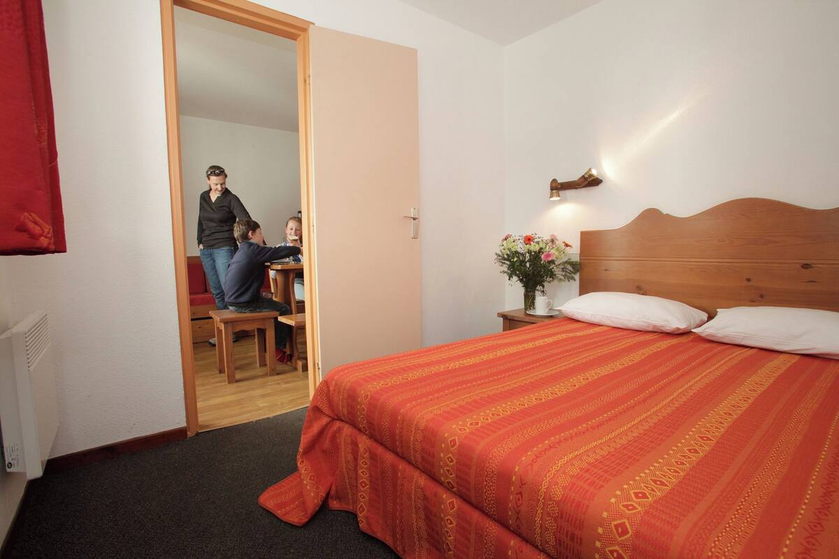 Ein Schlafzimmer mit hölzernem Kopfteil, einer roten, gemusterten Bettdecke, einem Nachttisch mit Lampe und einer Tür zu einem anderen Raum.