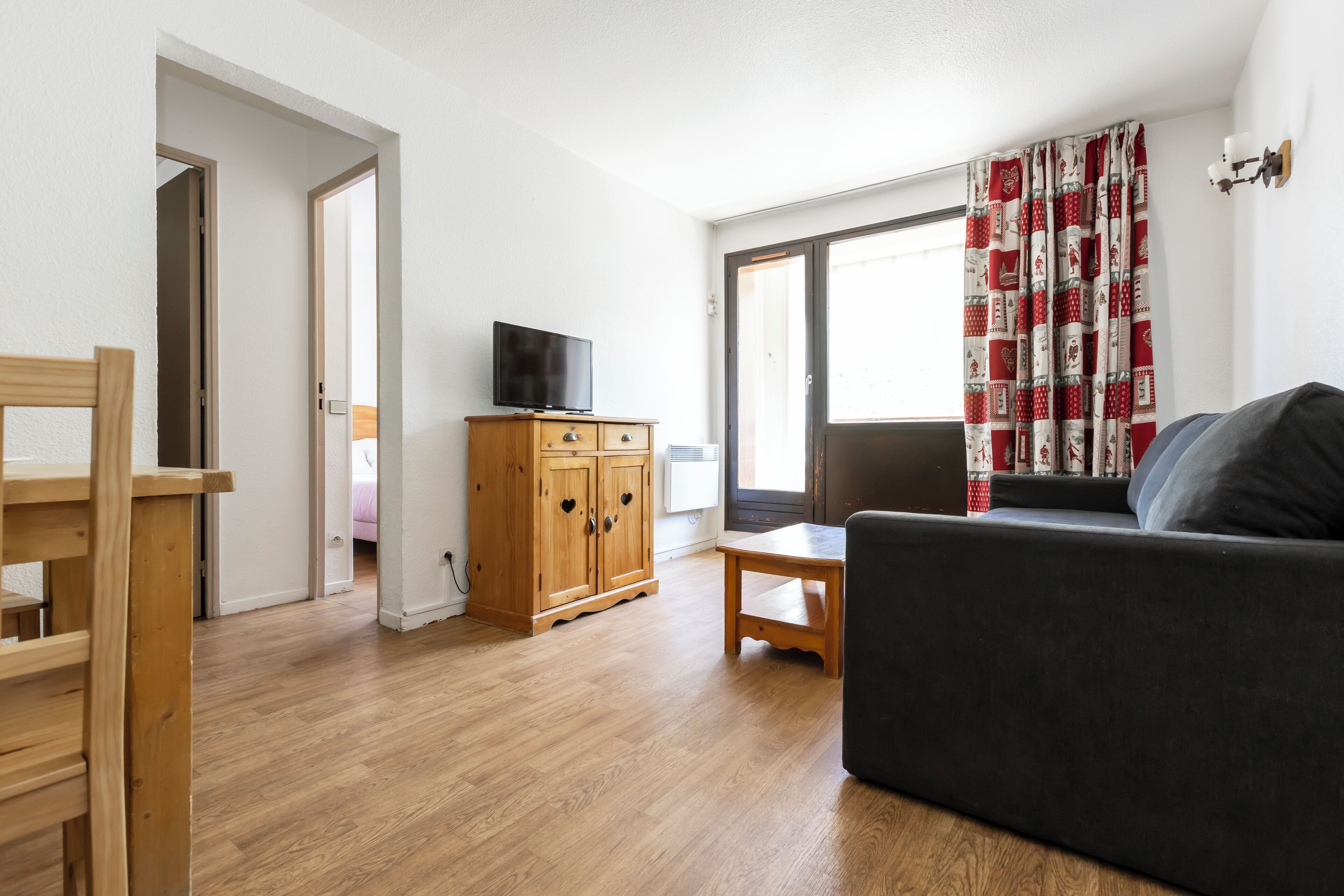Ein Wohnzimmer mit einem Holzkabinett, einem Fernseher, einer Couch, einem Tisch und Blick durch ein Fenster mit Vorhängen.