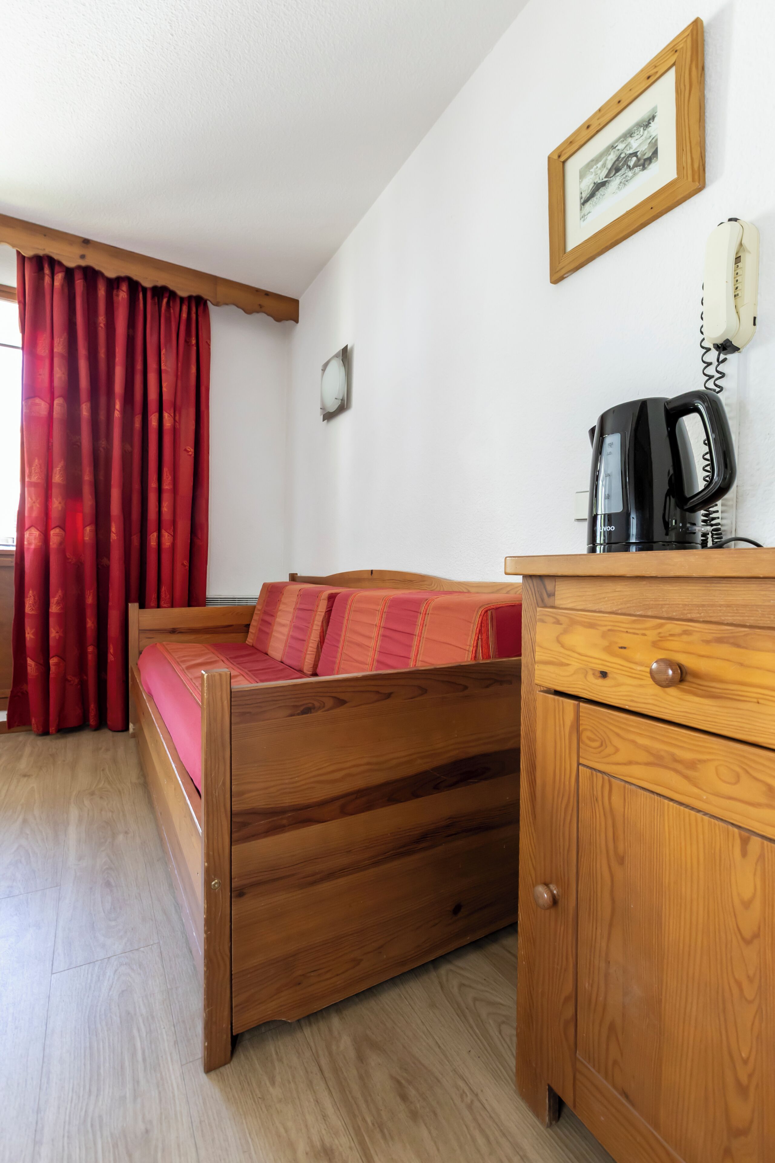 A wooden bed with a red blanket, a wooden nightstand with a coffee maker, a framed picture on the wall, and a red curtain.