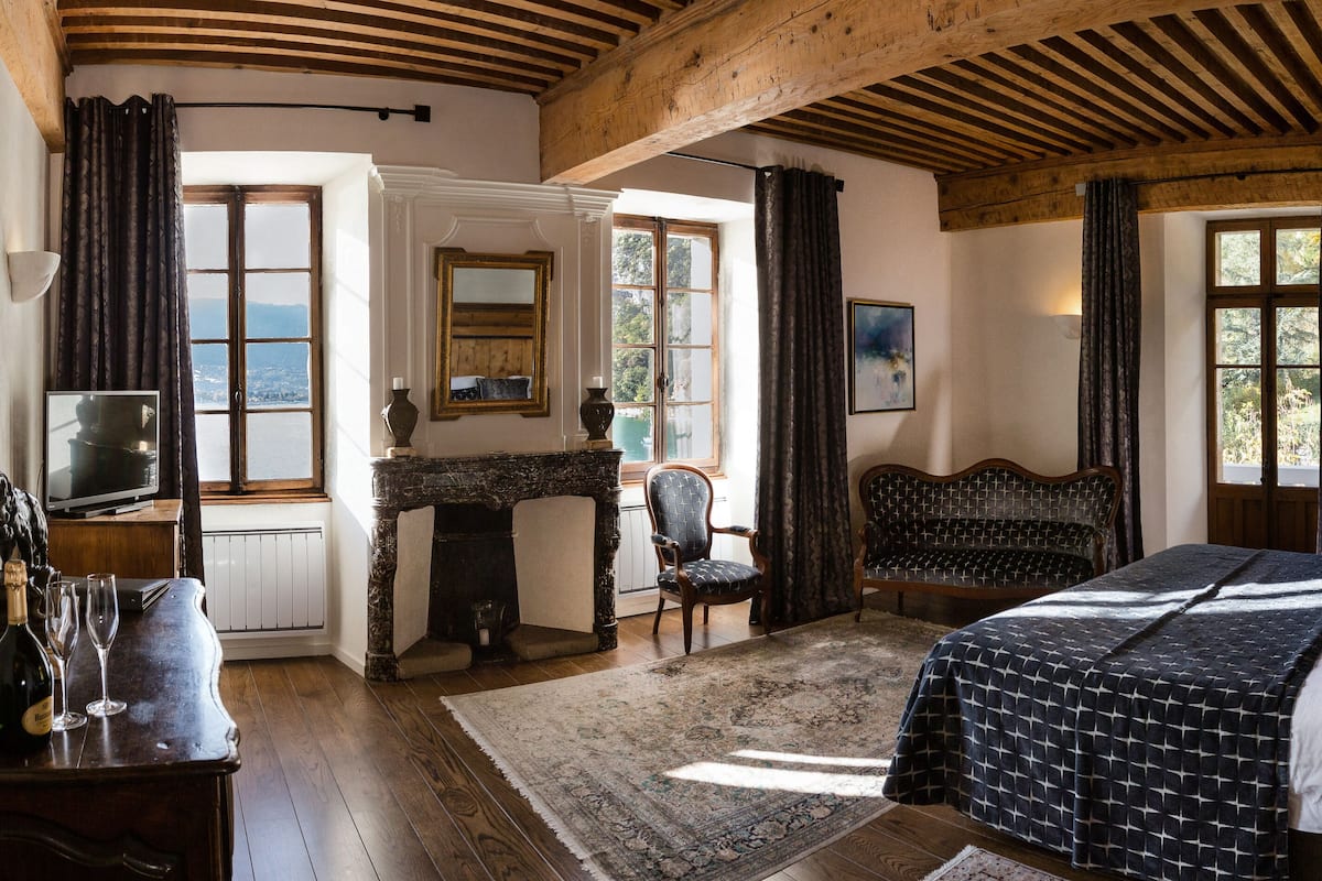A bedroom with a large bed, a fireplace, a wooden ceiling, and a view of the outdoors.