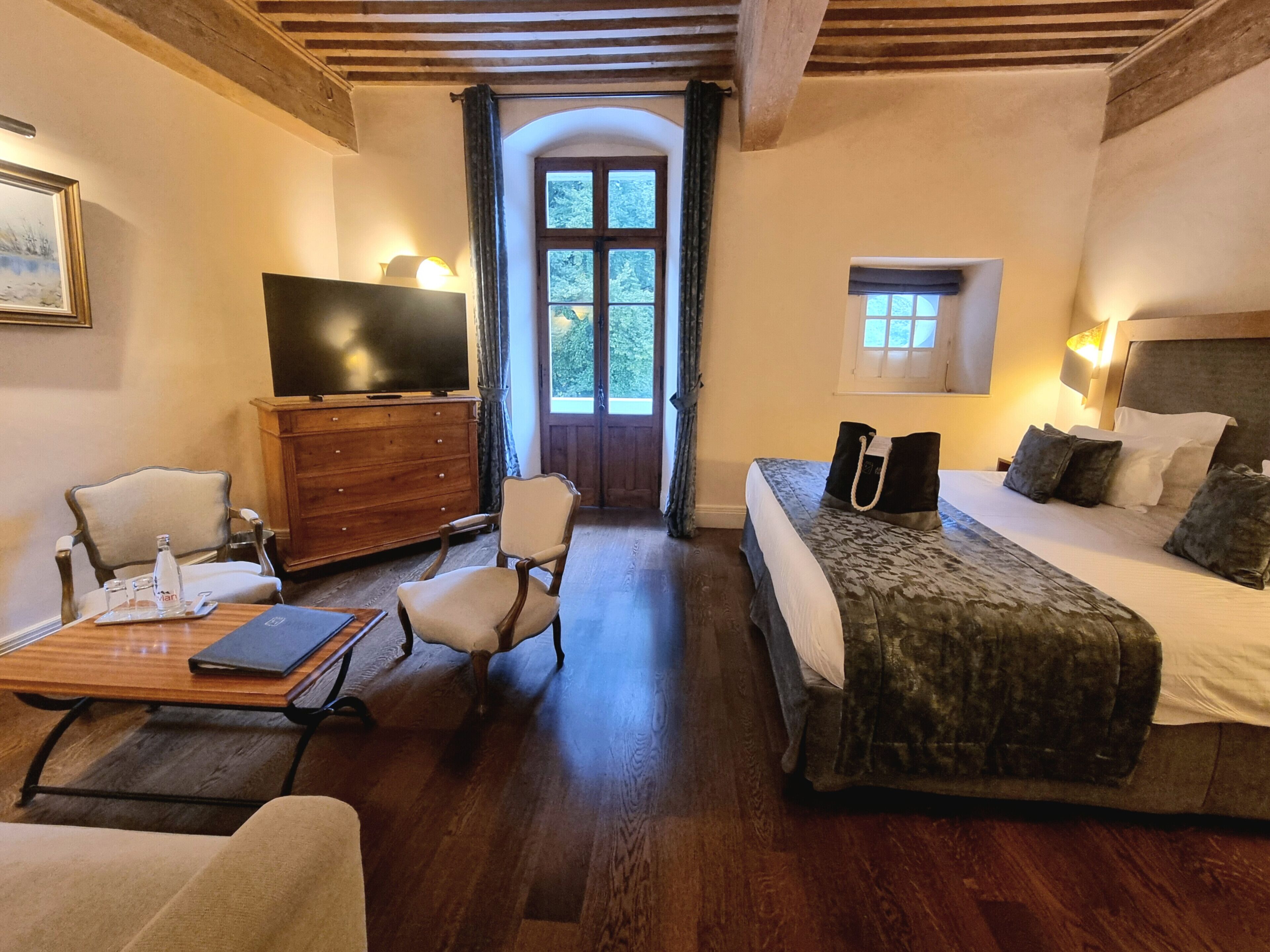 Quarto de hotel com piso de madeira, uma cama com colcha estampada, uma televisão sobre uma cômoda de madeira, uma cadeira, uma mesa pequena com bandeja e uma janela com cortinas.