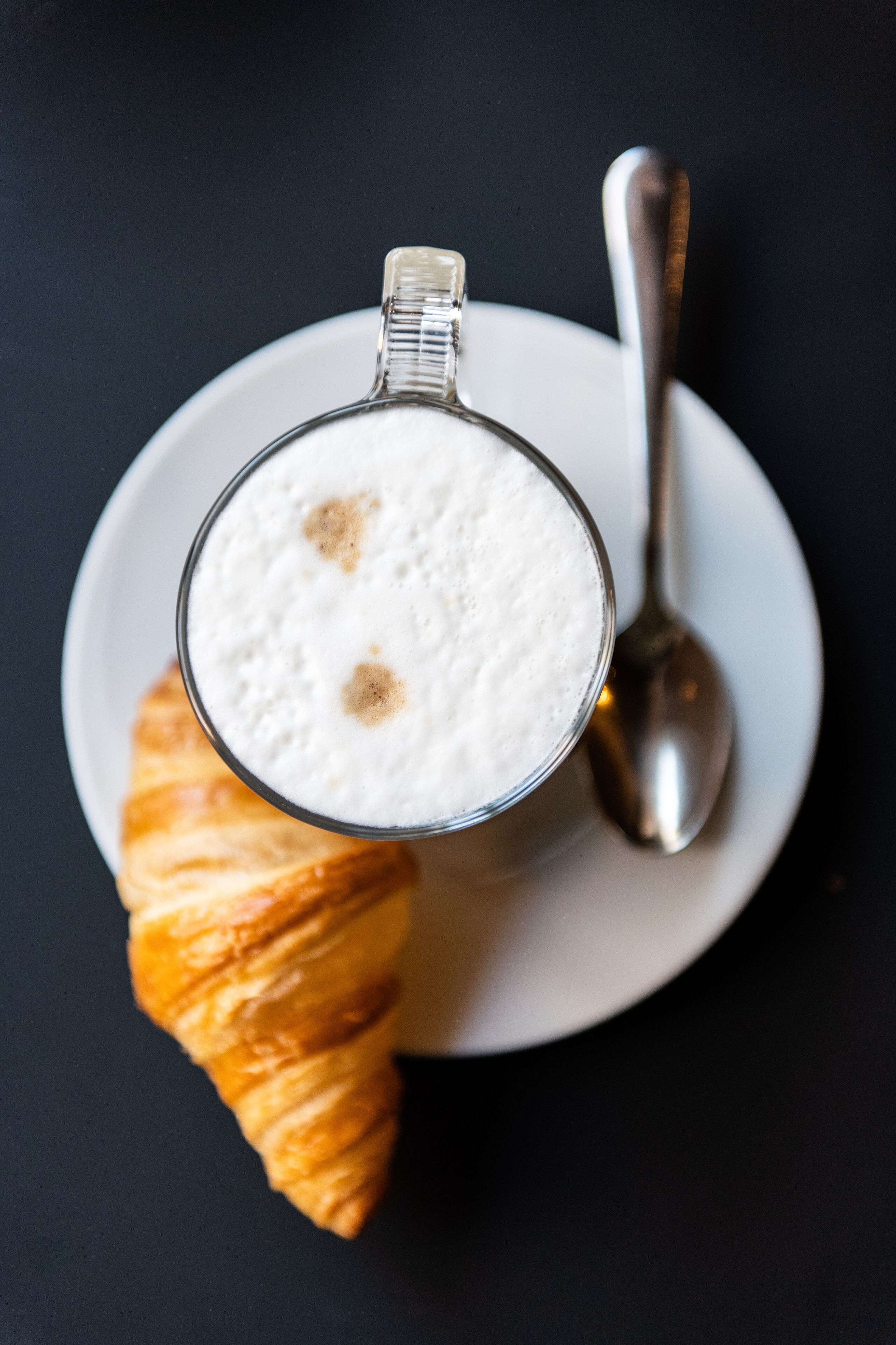 Una taza de capuchino con un croissant sobre un plato blanco, junto a una cuchara.