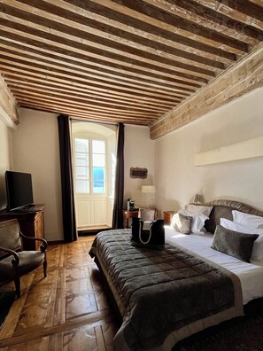 Habitación de hotel con techo de madera, cama con colcha oscura, escritorio con lámpara y vista por una ventana.