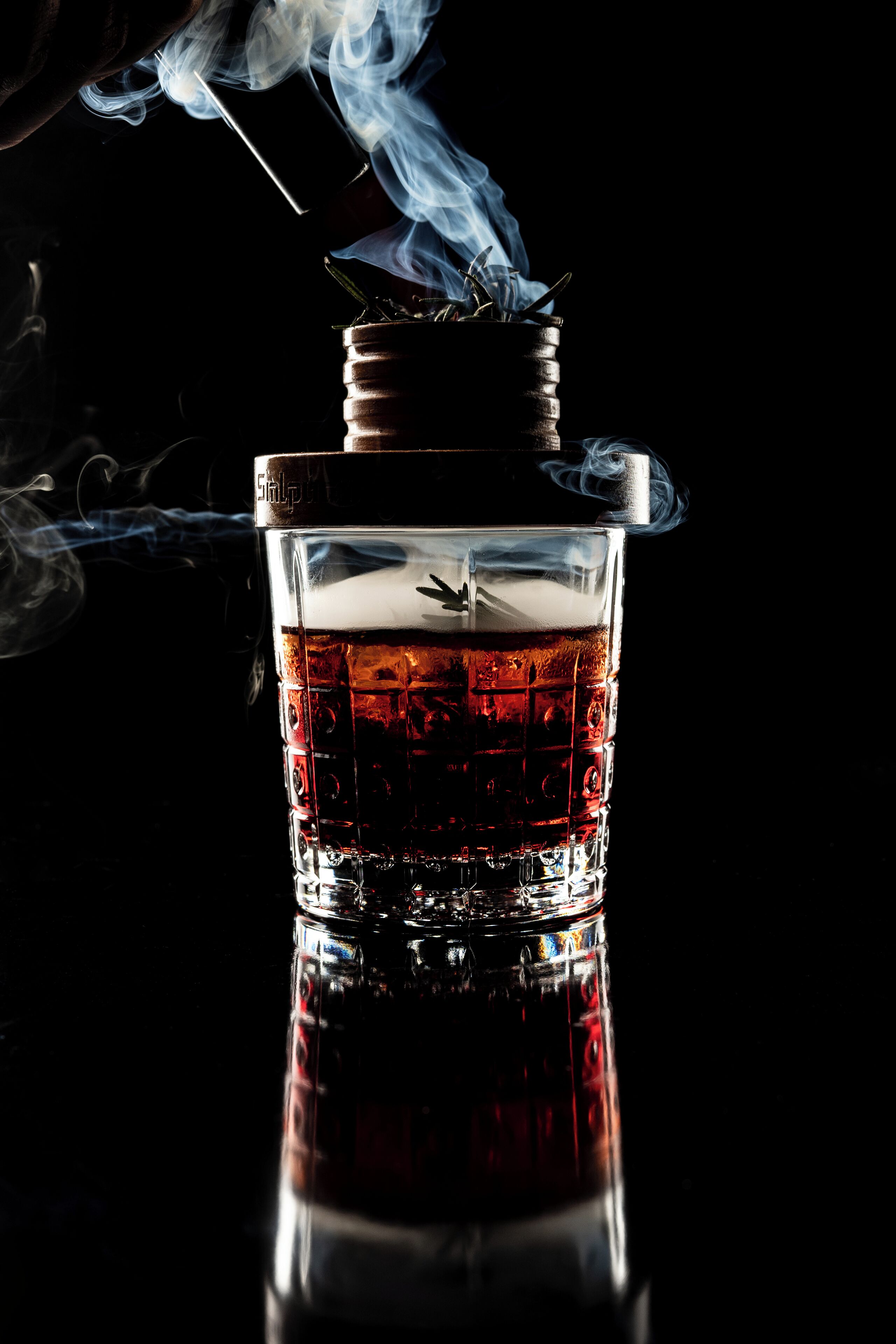 A glass of whiskey with a smoking glass on top, set against a dark background.