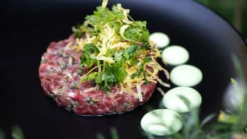 A steak tartare dish on a black plate with garnishes, surrounded by greenery.