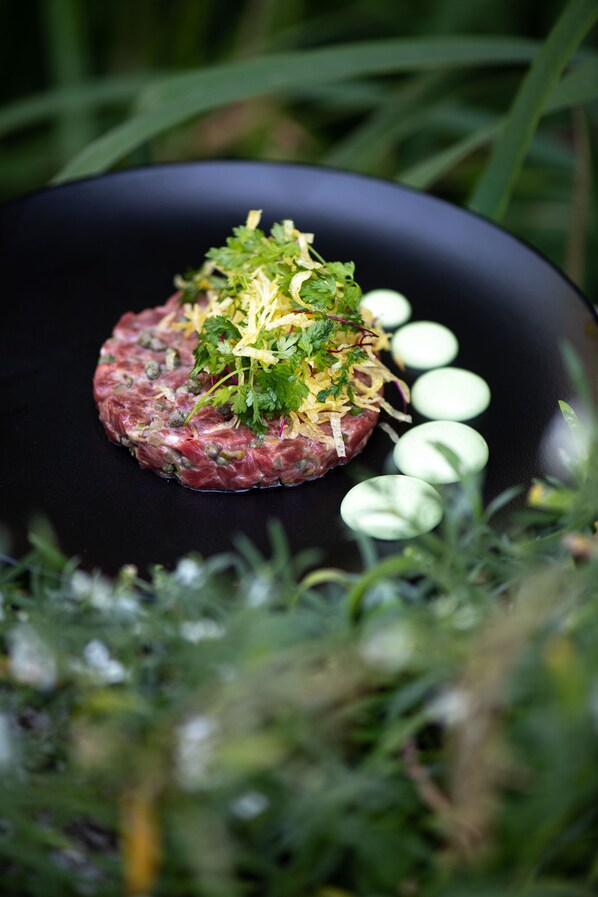 Un plat de steak tartare servi dans une assiette noire, agrémenté de quelques accompagnements et entouré de verdure.