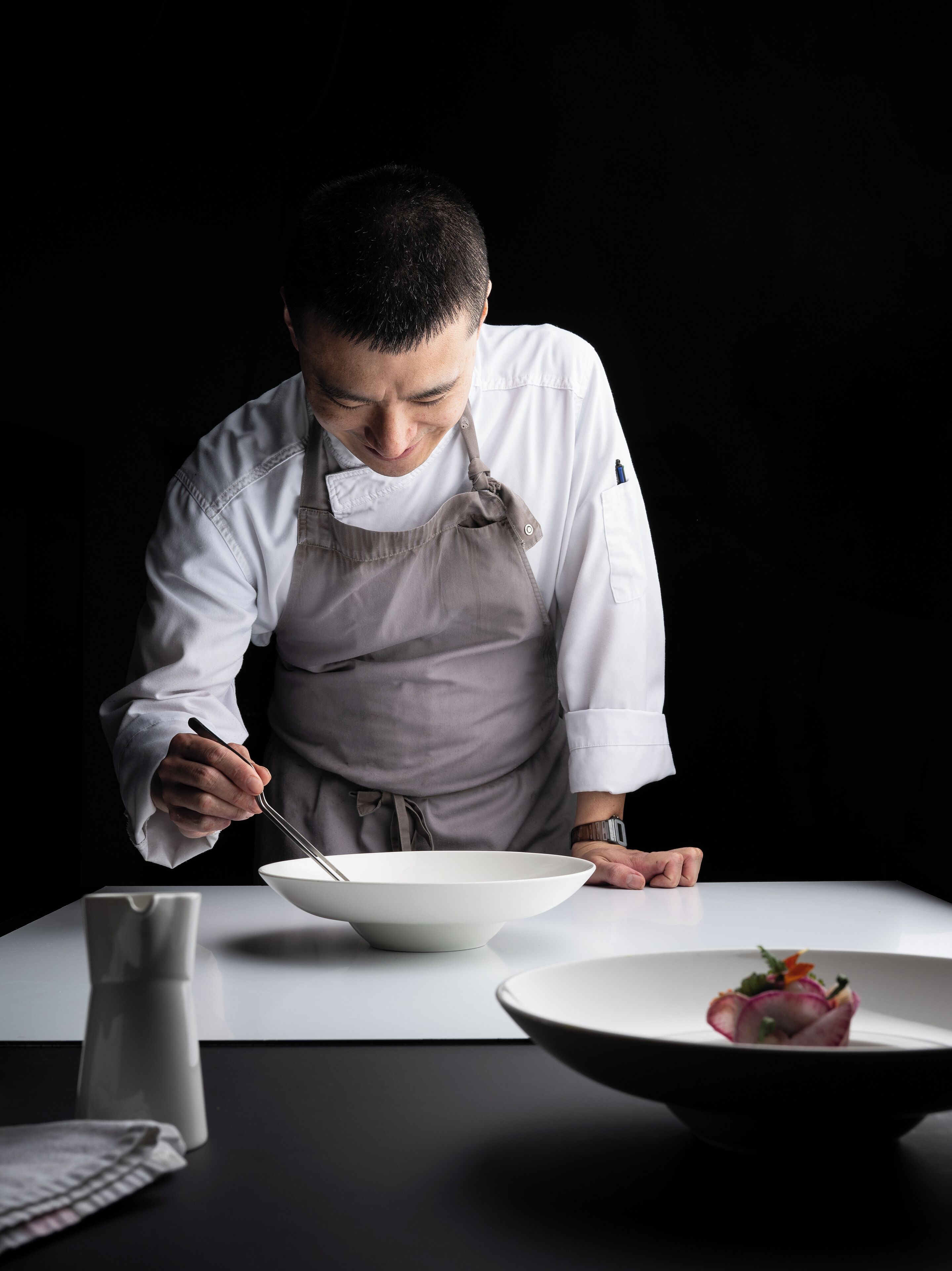 A chef in a white uniform with a gray apron, preparing food at a table with a white bowl and a plate with a dish.