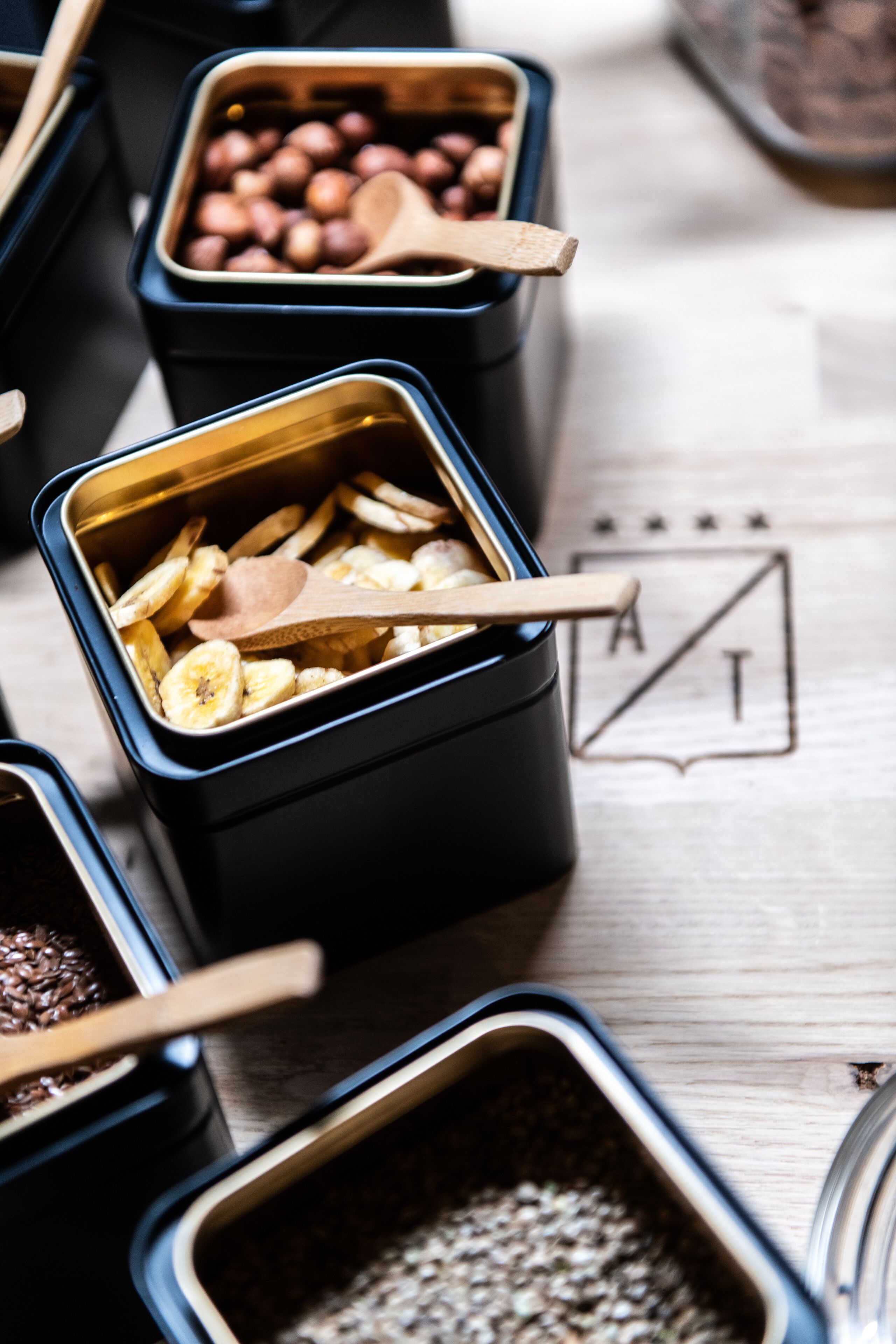 Multiple containers with wooden spoons, one containing sliced bananas and another with nuts.