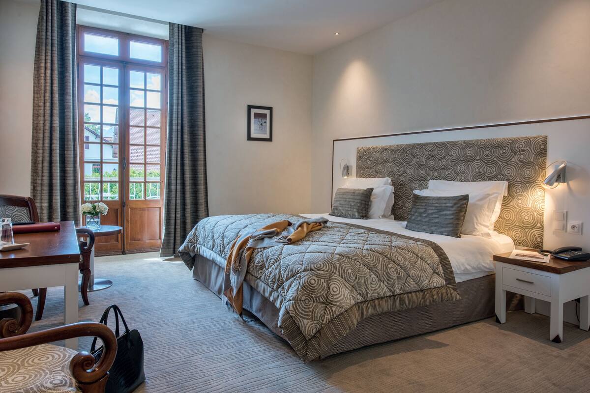 A hotel room with a large bed, a desk, a chair, a nightstand, and a window with curtains.