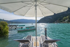 Terraza de madera con sillones de descanso y una sombrilla, con vista a un lago tranquilo con barcos y montañas al fondo.