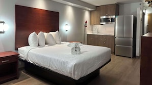 Habitación de hotel con cama, mesita de noche, luz empotrada en la pared, cabecera de madera, cocina integrada con microondas y refrigerador, y extintor.