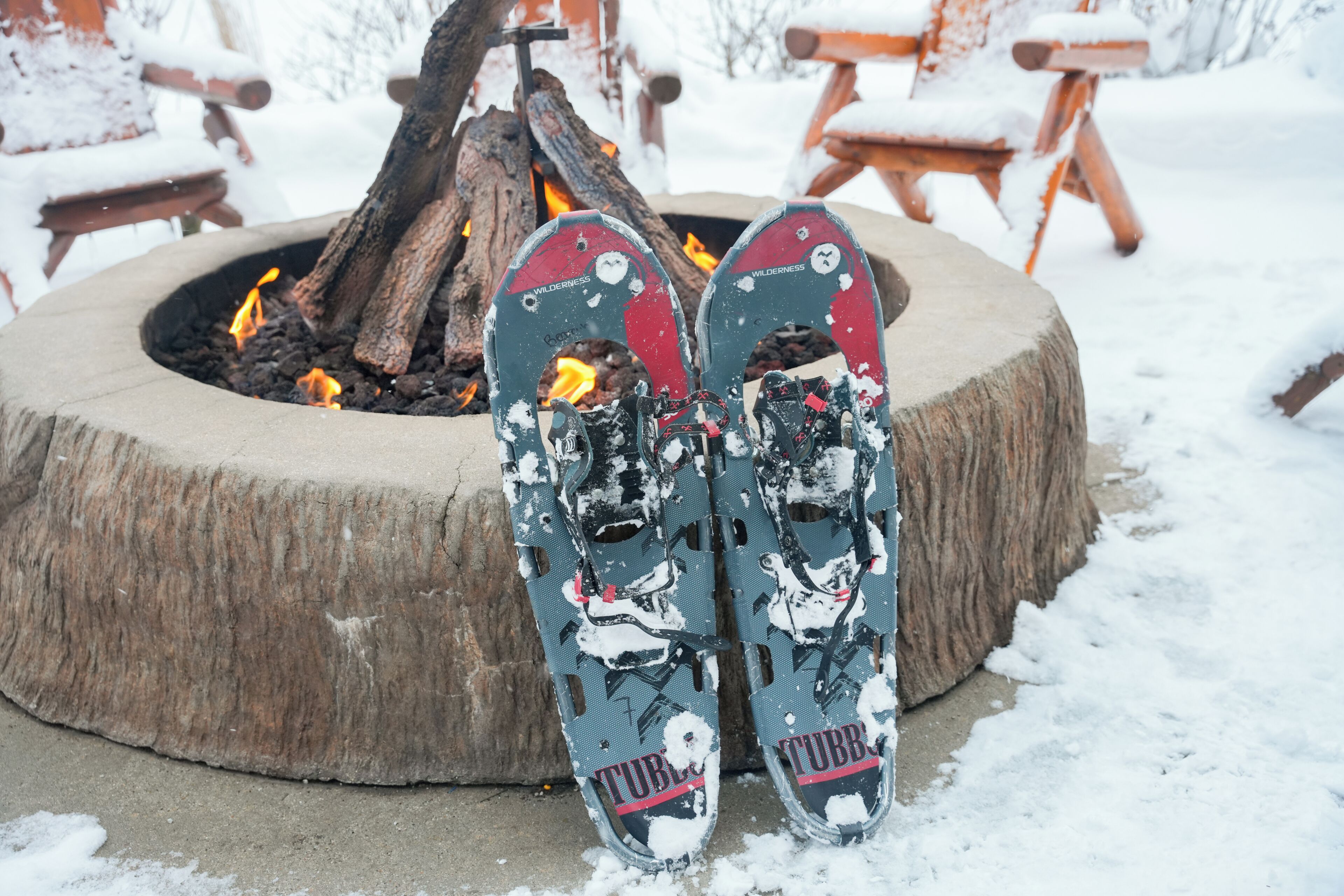 A pair of snowshoes with a 'Wilderness' logo and 'TUB' branding, placed in front of a fire pit with burning logs.
