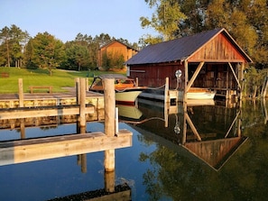Un muelle de madera con una estructura cubierta, un bote y un área verde con árboles al fondo.
